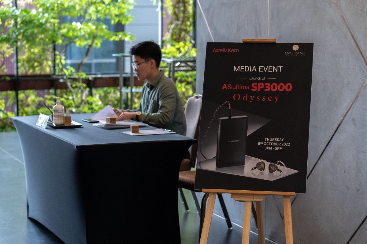 A large poster, featuring details of the Astell&Kern SP3000 and Odyssey media launch event, is displayed on a wooden easel stand near the registration table where a young man is seated to welcome the attendees.